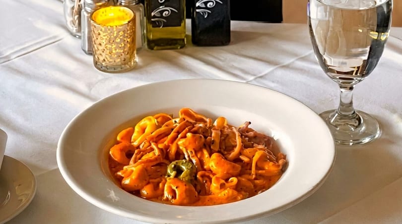 A white bowl of tortellini in a creamy orange sauce on a white tablecloth with a candle and water glass.