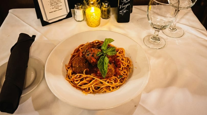 Spaghetti and meatballs with basil on a white plate, with wine glasses and a candlelit table.